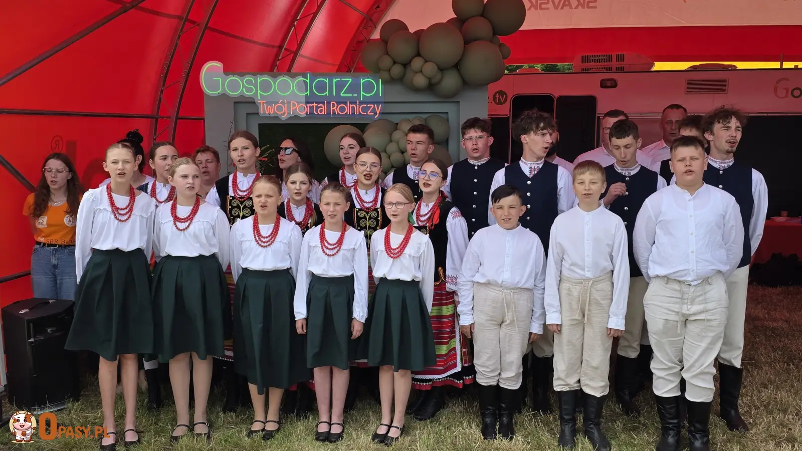 Zespół Pieśni i Tańca „Szepietowo na Ludowo” podczas występu na stoisku portalu rolniczego Gospodarz.pl w ramach XXXI Regionalnej Wystawy Zwierząt Hodowlanych w Szepietowie. Dzieci i młodzież ubrani w tradycyjne stroje ludowe śpiewają na tle oznakowanego stoiska.