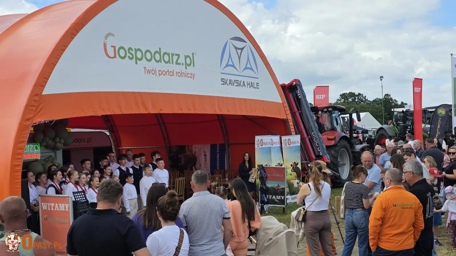 Stoisko Gospodarz.pl podczas XXXI Regionalnej Wystawy Zwierząt Hodowlanych w Szepietowie, przed którym zgromadził się tłum odwiedzających, oglądających występ artystyczny młodzieży; w tle widać maszyny rolnicze oraz namioty innych wystawców. 